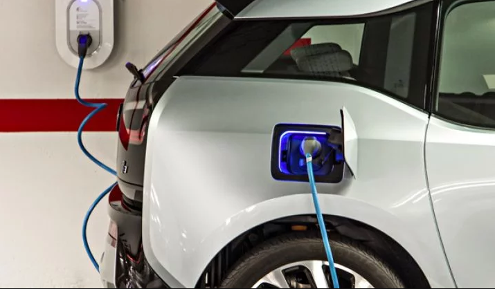 Please create a brief 1 sentence description of the attached imageEditThe image shows a white electric vehicle being charged in a garage, with a blue charging cable connected from a wall-mounted charging station to the car's illuminated charging port.