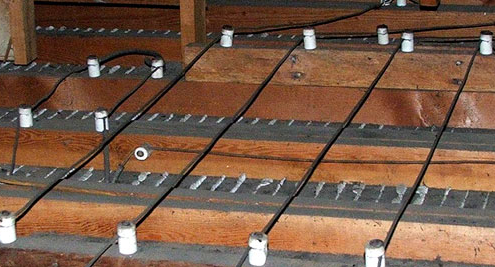 The image shows wooden floor joists or framing members with electrical wiring running through them, secured by white cable clamps or clips for residential electrical installation.