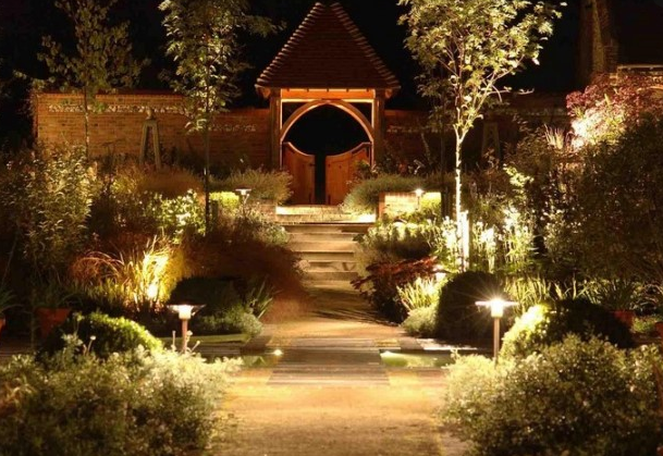 The image shows an elegantly lit garden pathway at night leading to a wooden gazebo or pavilion, with landscape lighting illuminating the walkway, surrounding plants, and architectural features.